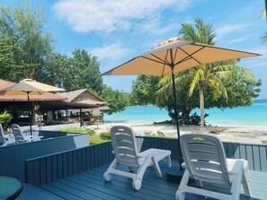 Une terrasse avec des chaises longues et une table, donnant sur une plage aux eaux bleu cristallin et bordée de palmiers.