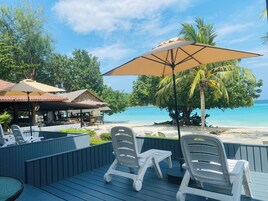 A deck with lounge chairs and a table, overlooking a beach with clear blue water and palm trees.