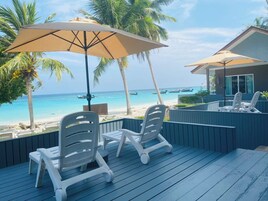 A deck with white plastic chairs and a large beige umbrella, overlooking a beach with boats and palm trees.