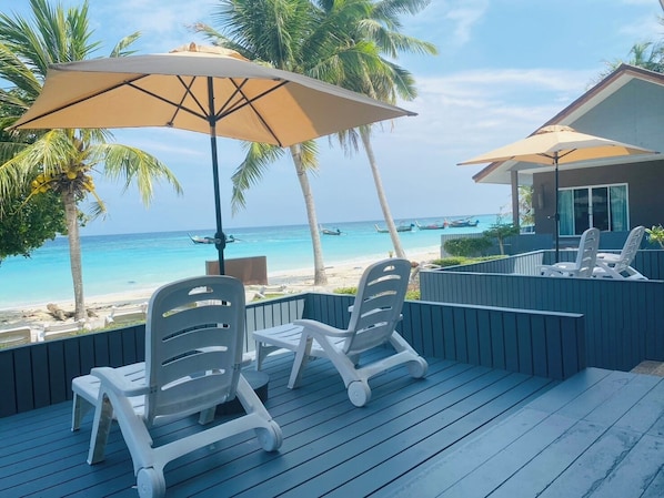 Eine Terrasse mit weißen Kunststoffstühlen und einem großen, beige farbenen Sonnenschirm, mit Blick auf einen Strand mit Booten und Palmen.