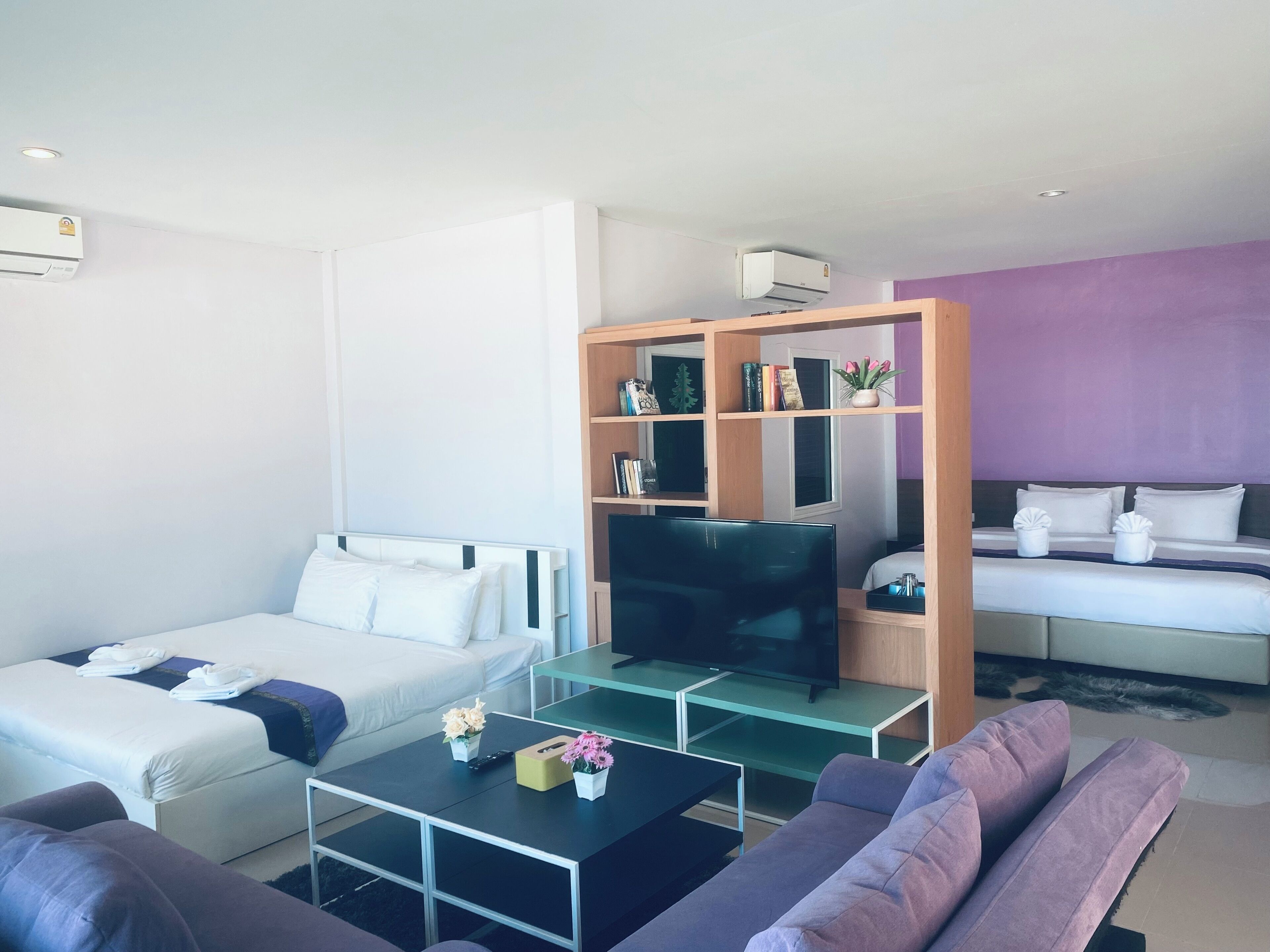 A modern hotel room with a sofa, a bed with white bedding, a television on a glass table, and a wooden bookshelf.
