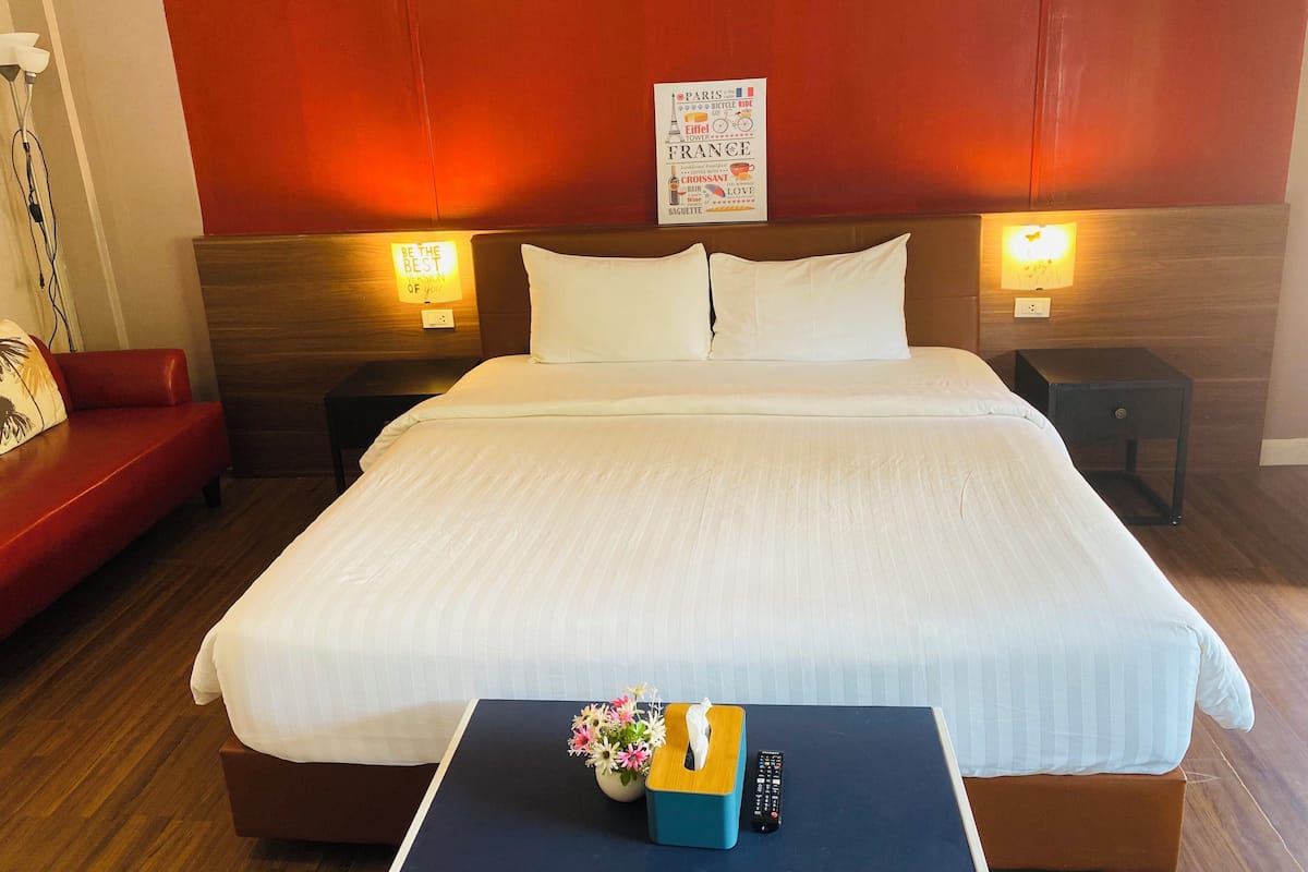Een hotelkamer met een groot bed, twee nachtkastjes met lampen, een rode bank en een kleine tafel met een afstandsbediening en een zakdoos.