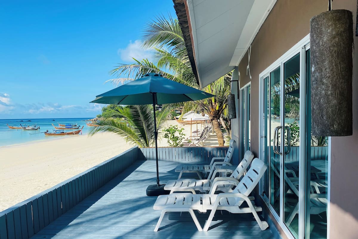 Een balkon aan het strand met witte plastic stoelen en een blauwe parasol, met uitzicht op een zandstrand met boten en palmbomen.