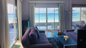 Een woonkamer met een paarse bank, een televisie en een salontafel. Er is een groot raam met uitzicht op het strand en de zee.
