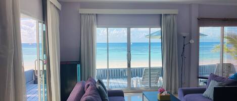 A living room with a purple sofa, a TV, and a coffee table. There is a large window with a view of the beach and ocean.