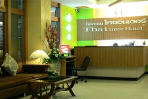 A reception area with a wooden reception desk, a green accent wall, and a sofa with a lamp and flowers. - Thai Inter Hotel (Nakhon Ratchasima)
