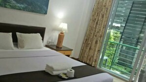 Habitación de hotel con cama, mesita de noche con lámpara, un cuadro en la pared y vistas a la vegetación a través de una ventana.