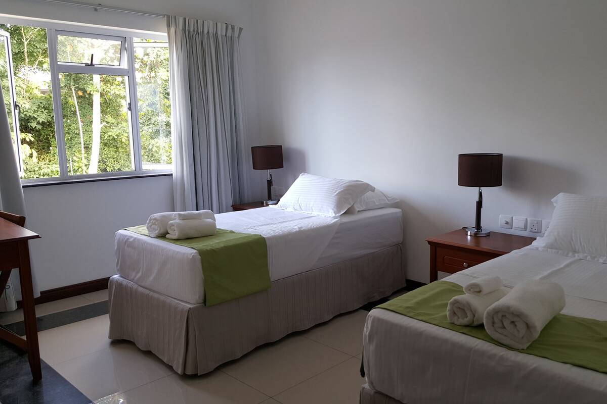 Habitación de hotel con dos camas, una ventana con cortinas y una mesita de noche con lámpara, además de un plaid verde sobre cada cama.