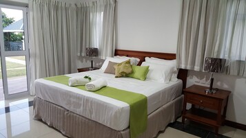 Una cama doble impecablemente hecha, con ropa de cama blanca y verde, un cabecero de madera y una mesita de noche con una lámpara.