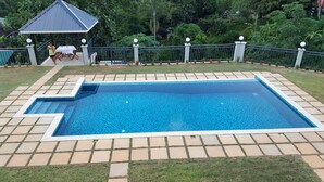 Ein rechteckiger Swimmingpool mit Fliesen, umgeben von einer Umzäunung und üppiger Vegetation.