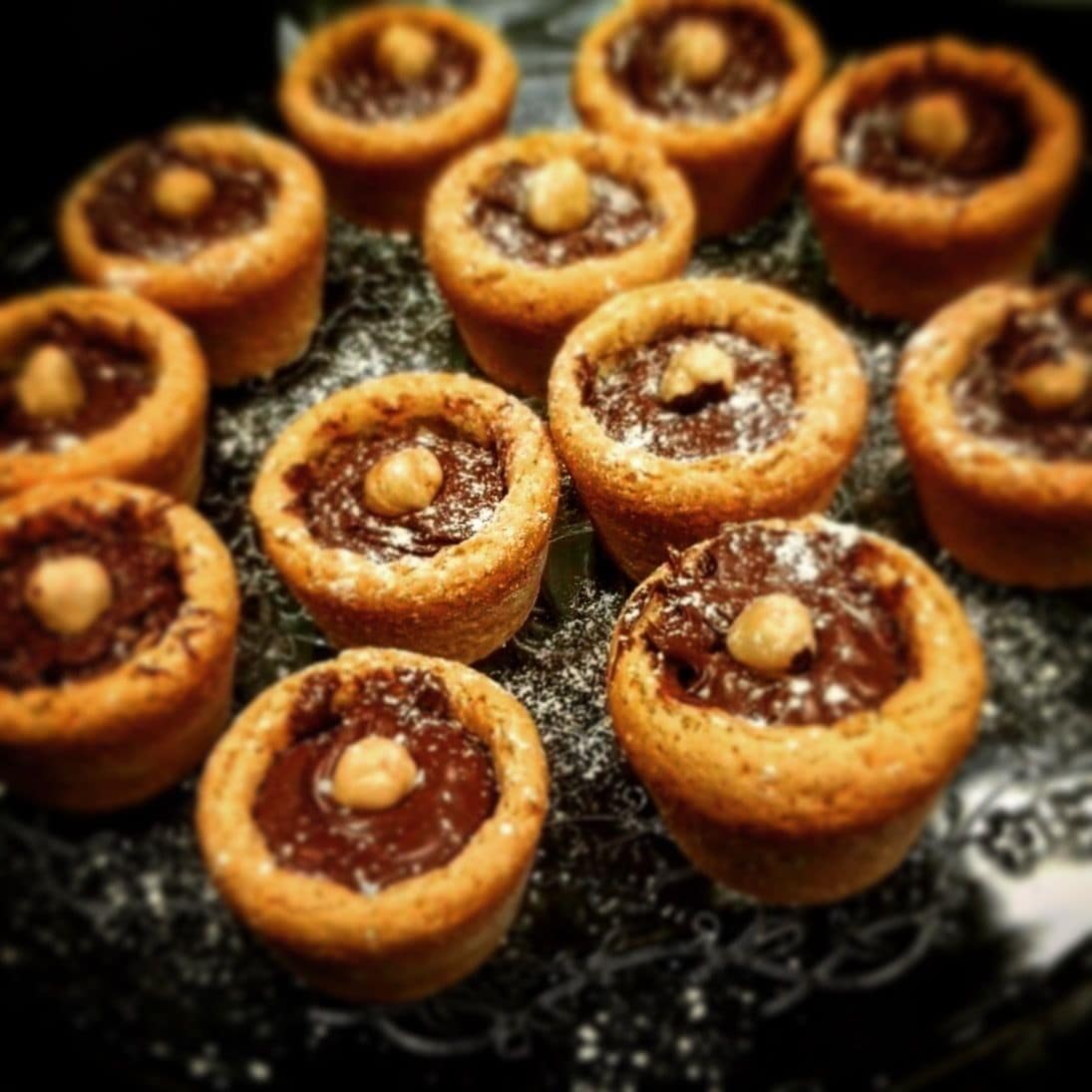Eine Schale mit Mini-Gebäckstücken, die goldbraune Kruste und dunkle Füllung haben und mit Puderzucker bestäubt sind.
