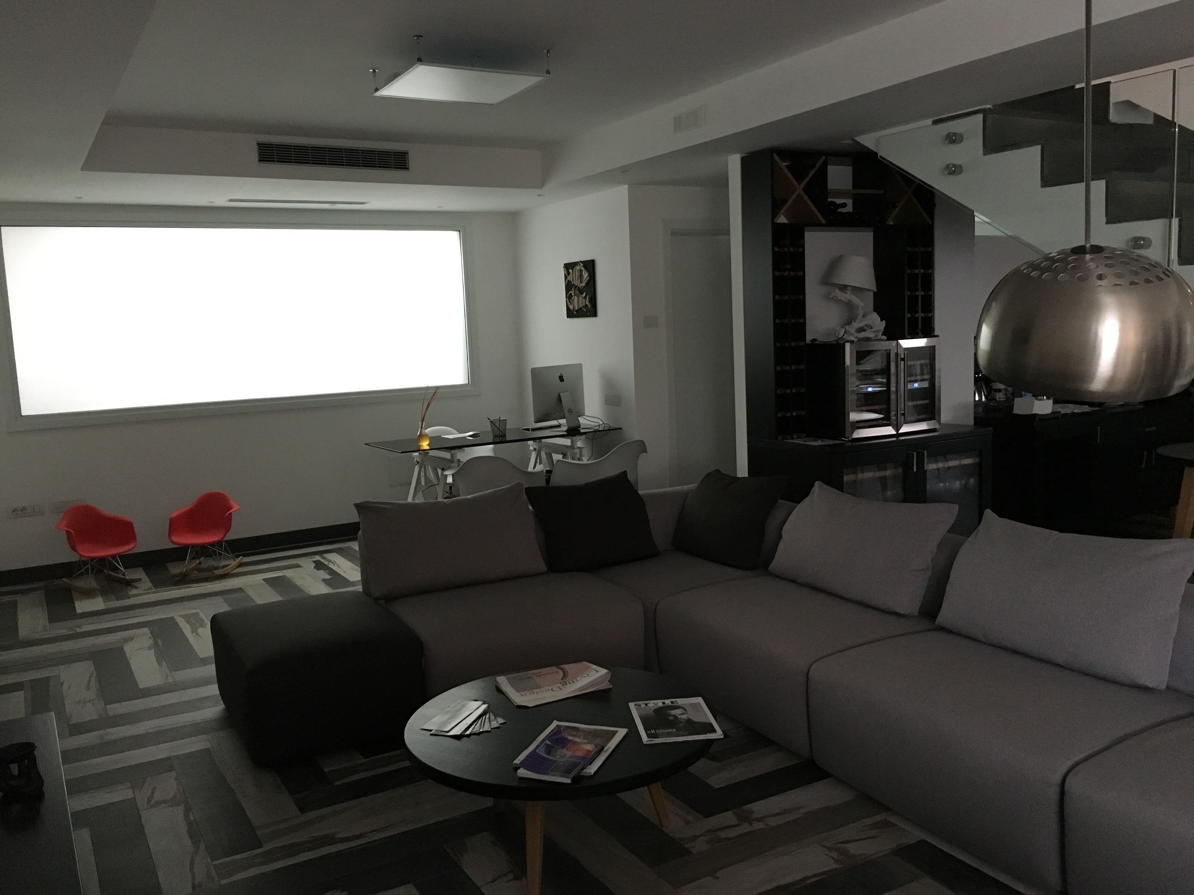 A modern living room with a grey sofa, a small round table with magazines, a red chair, and a large window providing natural light.