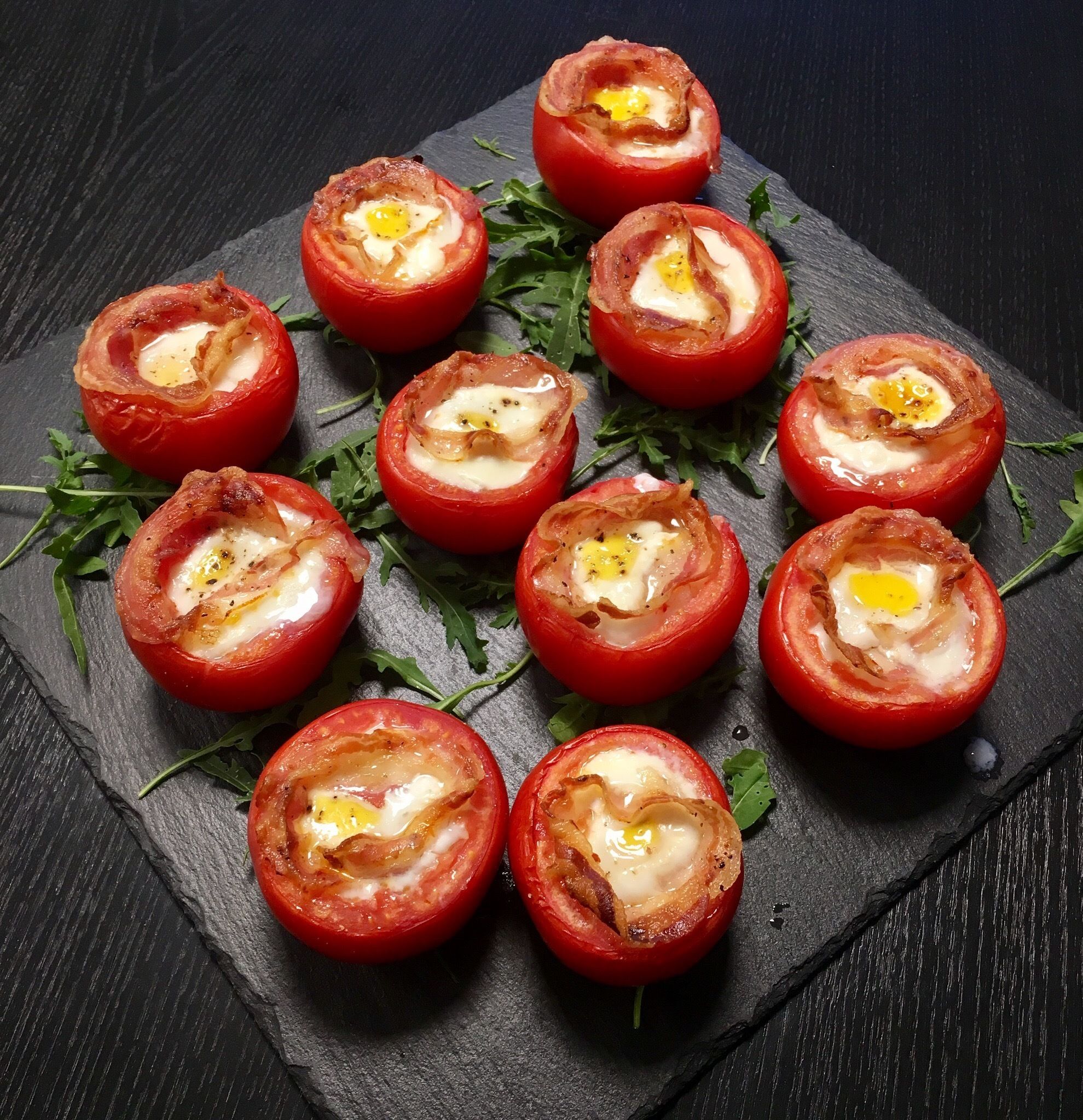 Una bandeja de tomates rellenos con un huevo escalfado, adornada con hierbas, sobre una tabla de pizarra.