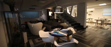 A modern living room with a dining area, featuring a sofa, chairs, a table, and a staircase leading to another level.