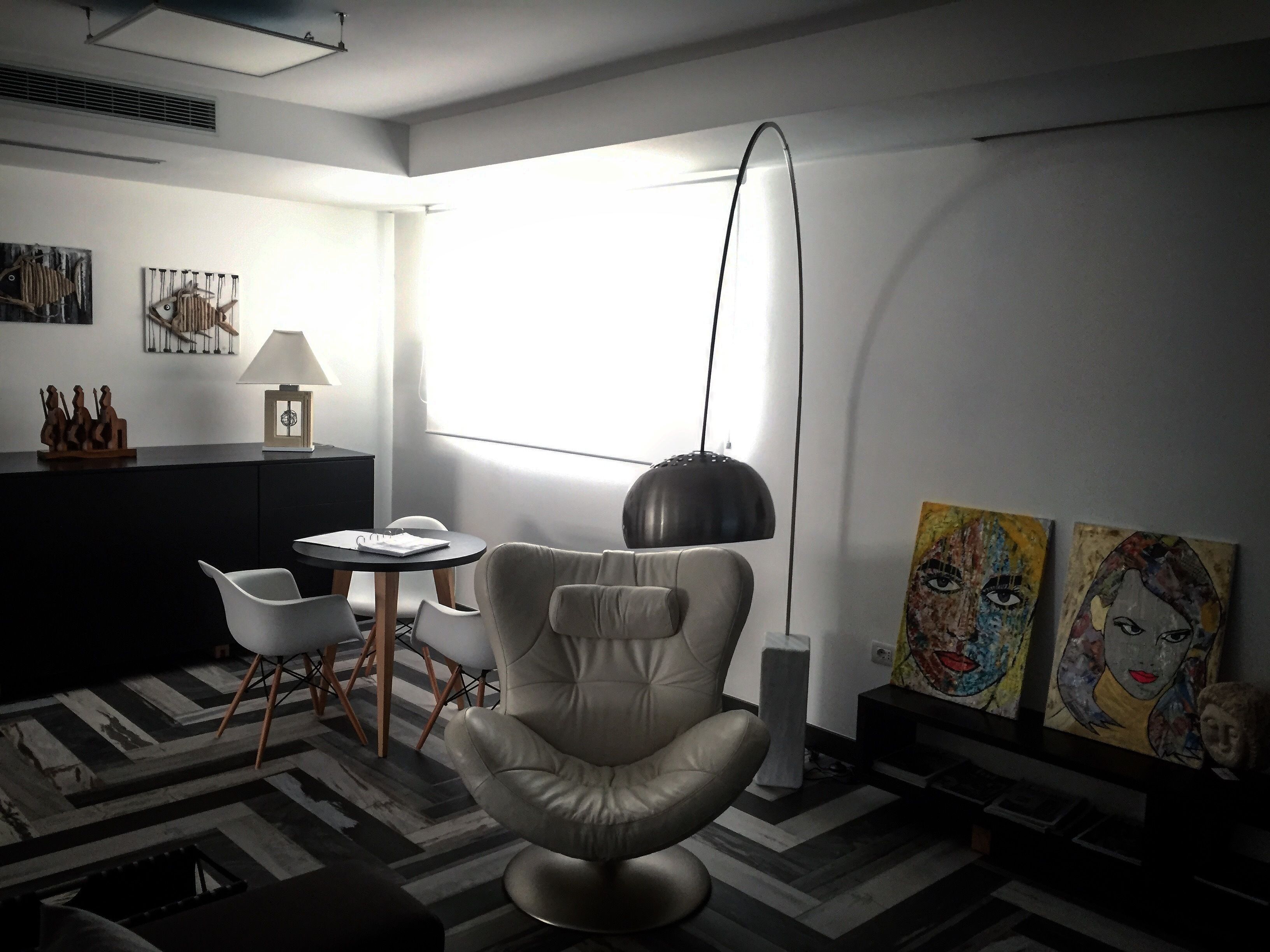 Uma sala de estar moderna com uma poltrona de couro, uma mesa redonda e um candeeiro pendente preto. Há duas pinturas na parede e uma janela que permite a entrada de luz natural.