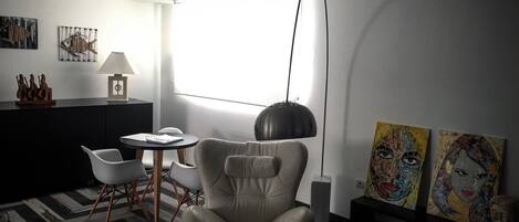 A modern living room with a leather armchair, a round table, and a black pendant lamp. There are two paintings on the wall and a window providing natural light.