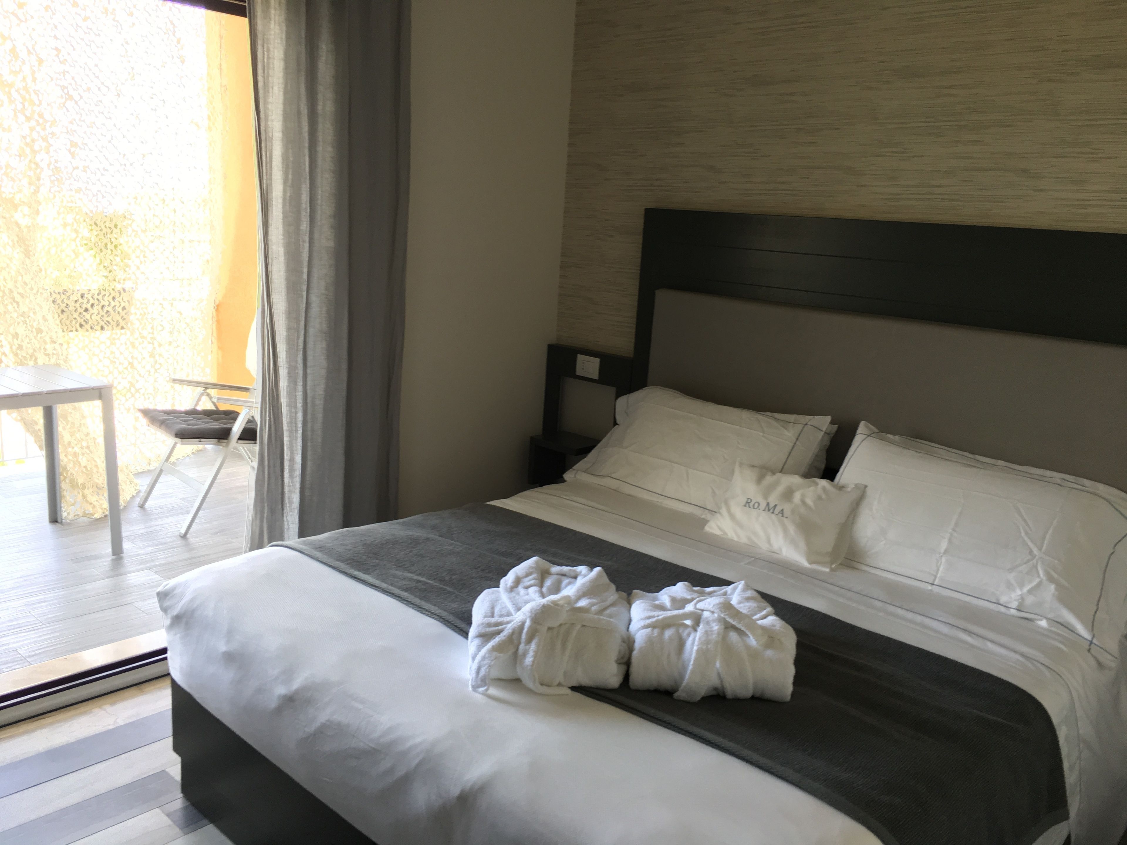 A neatly made bed with white linens and two towels, a small table, and a chair by a window with curtains.