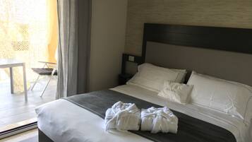 Una cama bien hecha con sábanas blancas y dos toallas, una mesita y una silla junto a una ventana con cortinas.