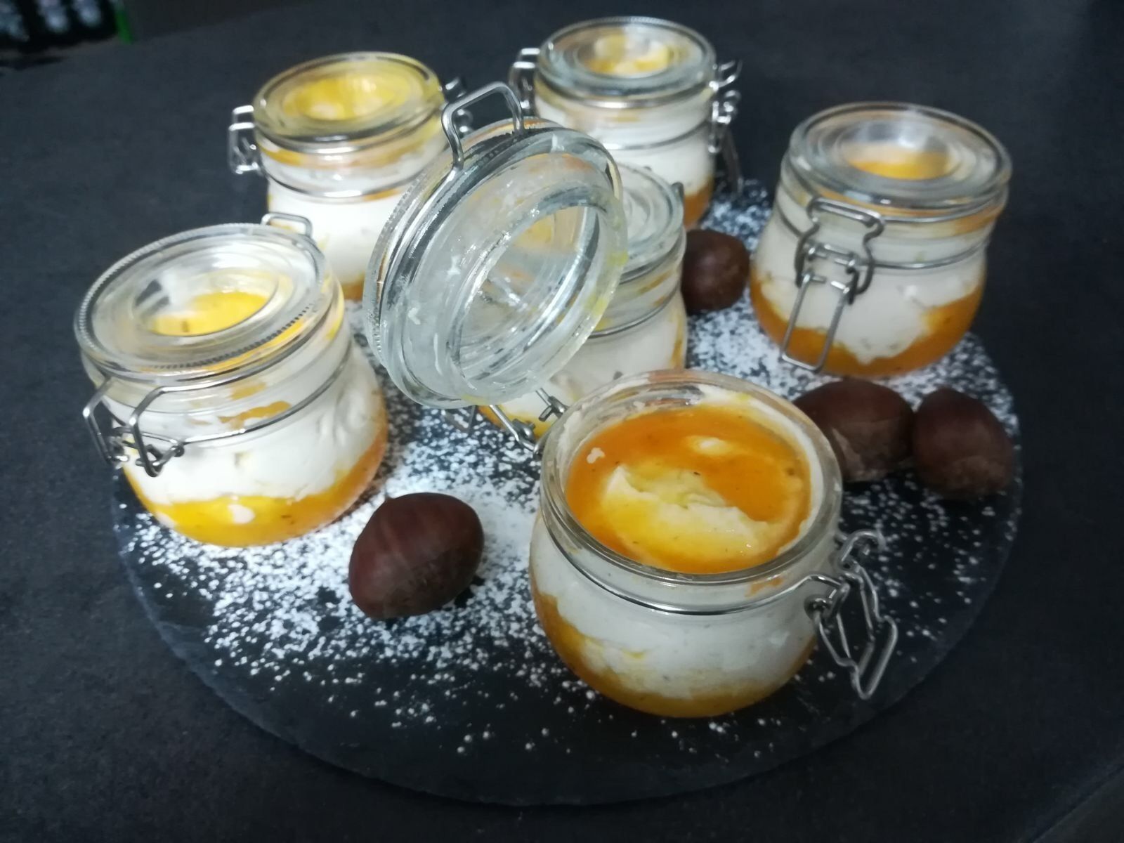 Six glass jars with metal clasps, each containing a layered dessert with a golden syrup topping, placed on a black slate board.