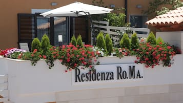 Ein Schild mit der Aufschrift 'Residenza Ro.Ma', eine weiße Wand, rote Blumen und ein weißer Sonnenschirm.
