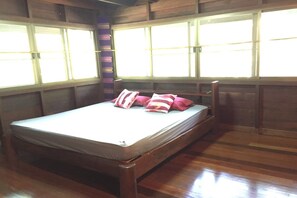 A single bed with a wooden frame and a mattress, positioned against a wall with large windows. - Baan Parimtarn Kanchanaburi (Kanchanaburi)