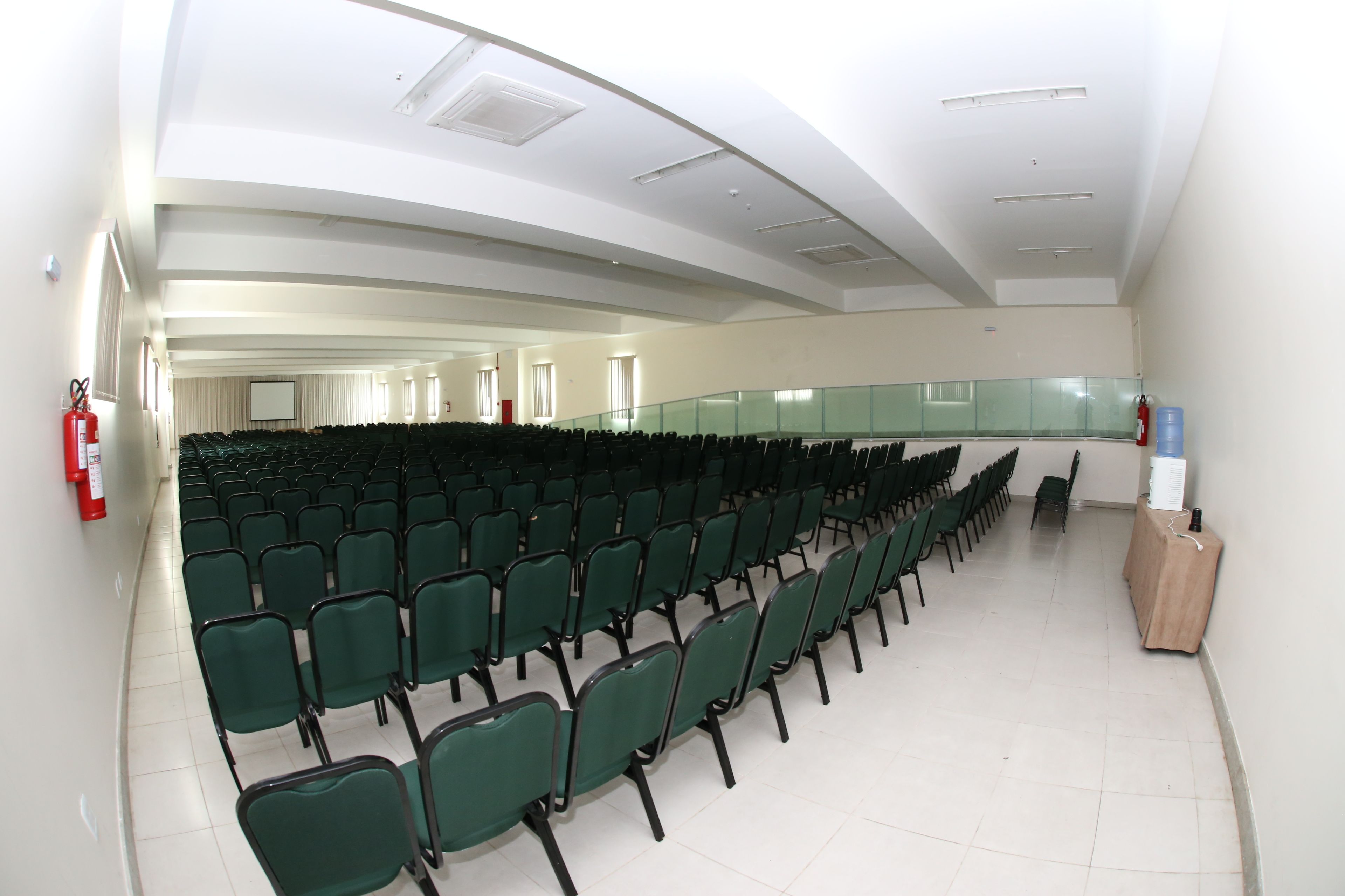 Sala de conferência com fileiras de cadeiras verdes, teto branco e extintor de incêndio à esquerda.