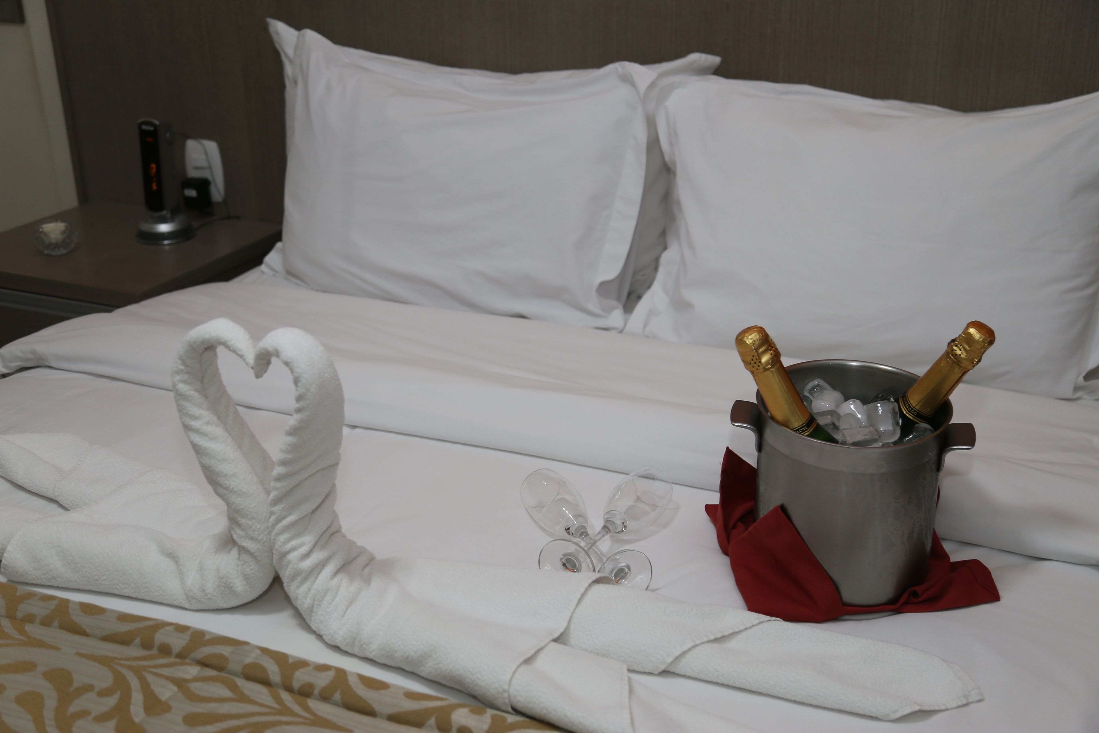 A bed with white linens, a heart-shaped towel, a bucket with champagne bottles, and a red napkin.