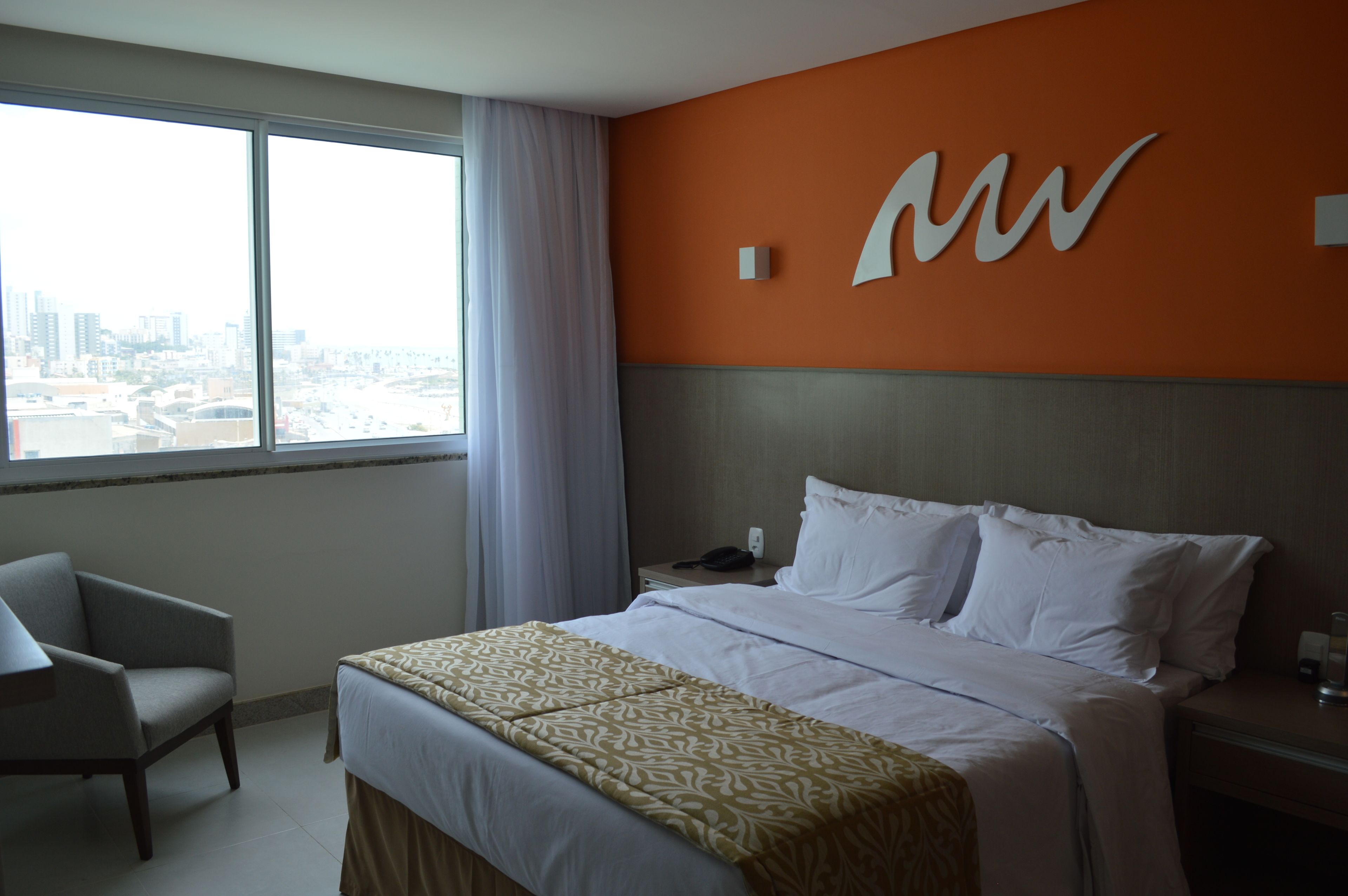 A hotel room with a large bed, a chair, a nightstand, and a window with a city view.