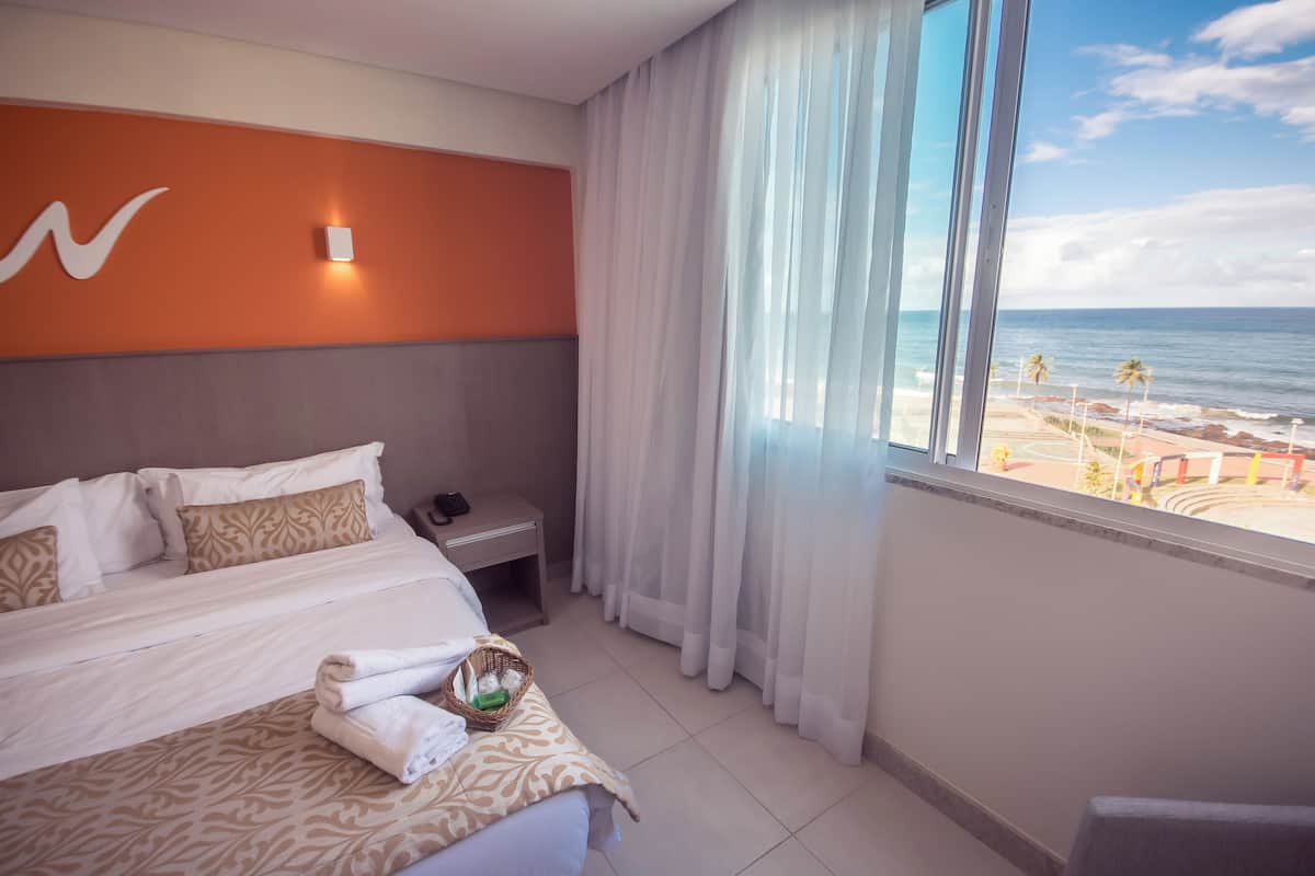 Een hotelkamer met een bed, een nachtkastje, een raam met uitzicht op het strand en een wand met een lamp.