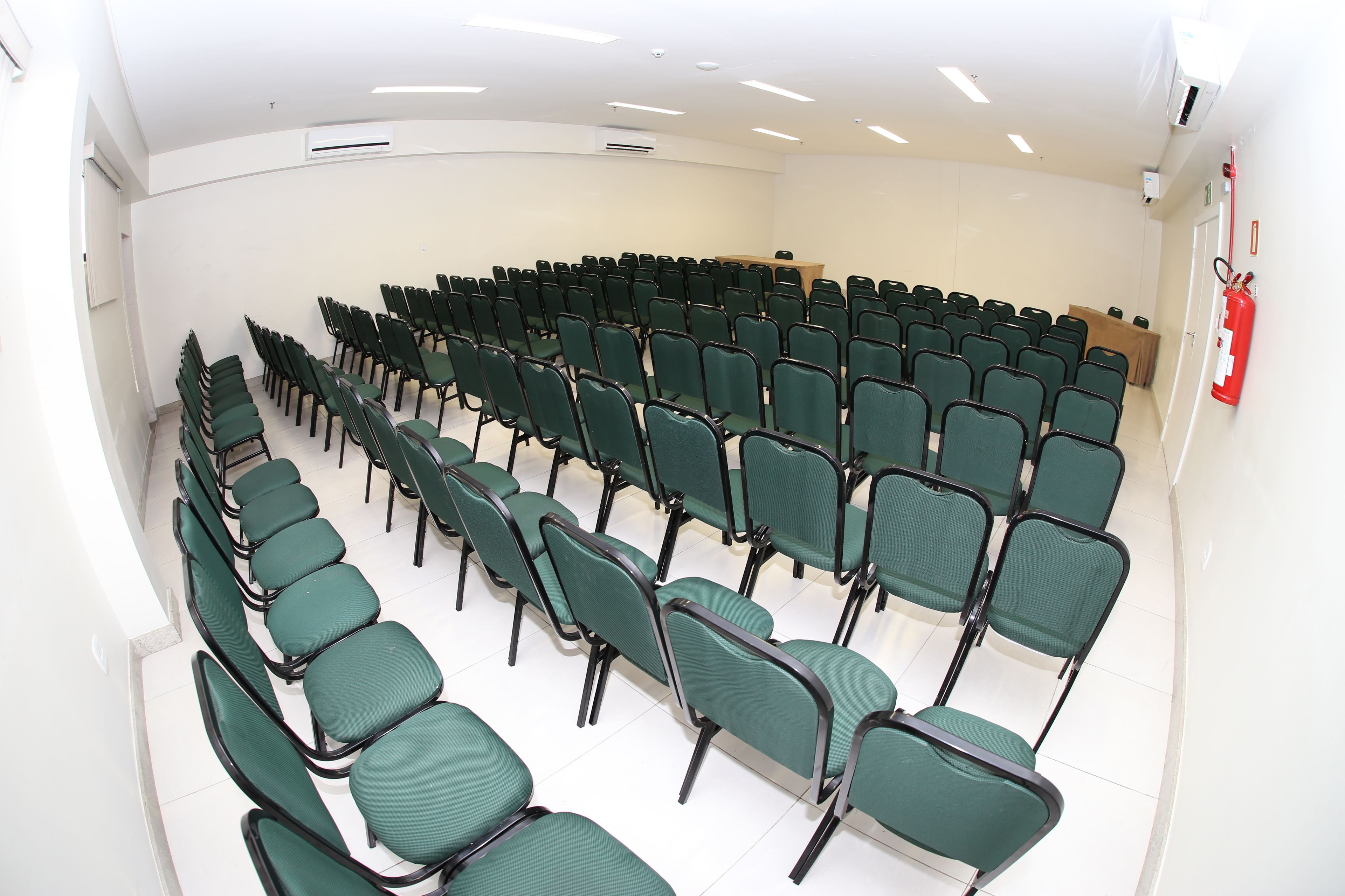 Uma sala de conferências com fileiras de cadeiras verdes, uma mesa de madeira e um extintor na parede.