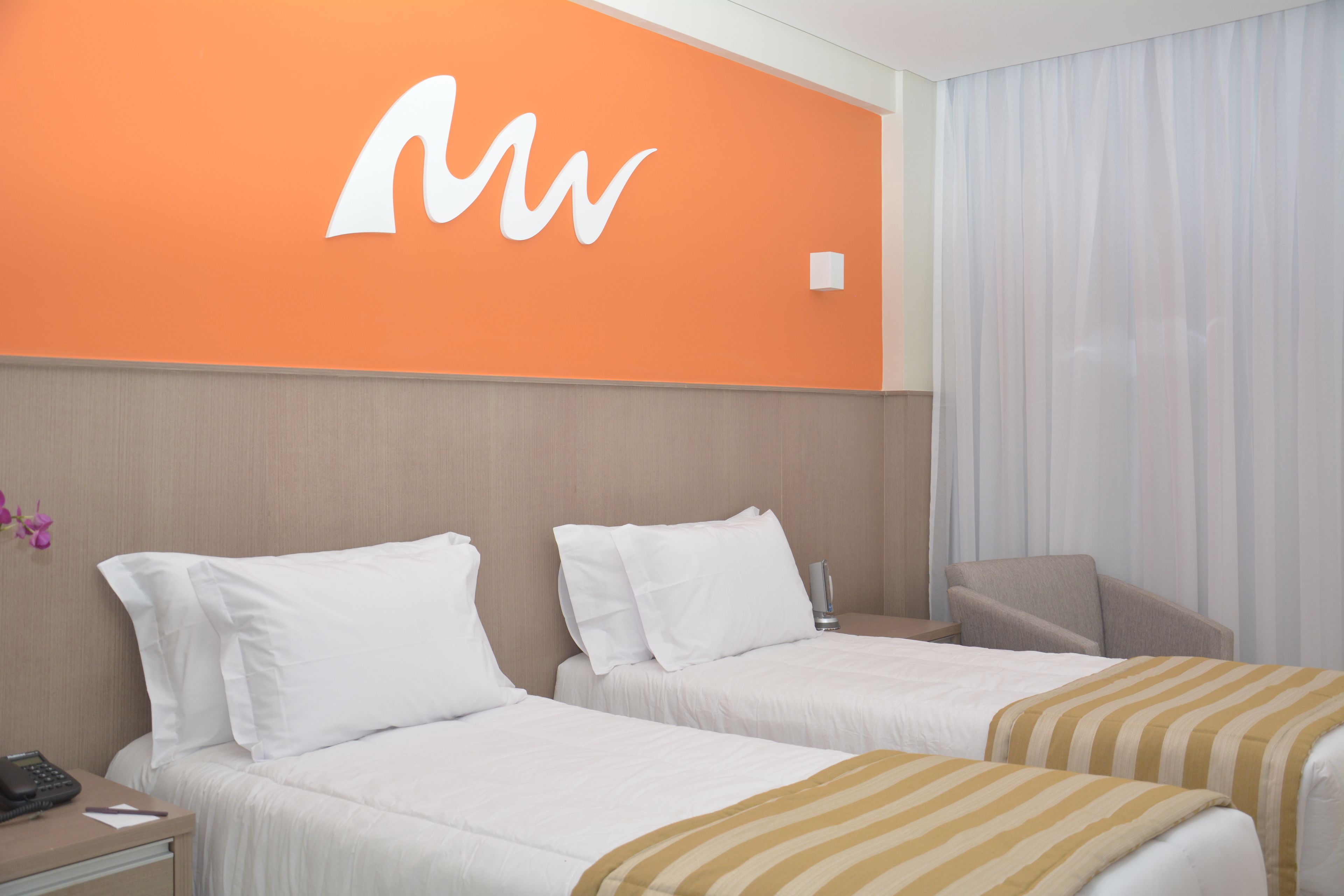 Quarto de hotel com duas camas, uma escrivaninha com telefone e uma parede laranja com logotipo branco.