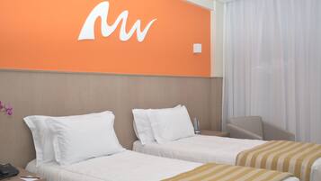 Quarto de hotel com duas camas, uma escrivaninha com telefone e uma parede laranja com logotipo branco.