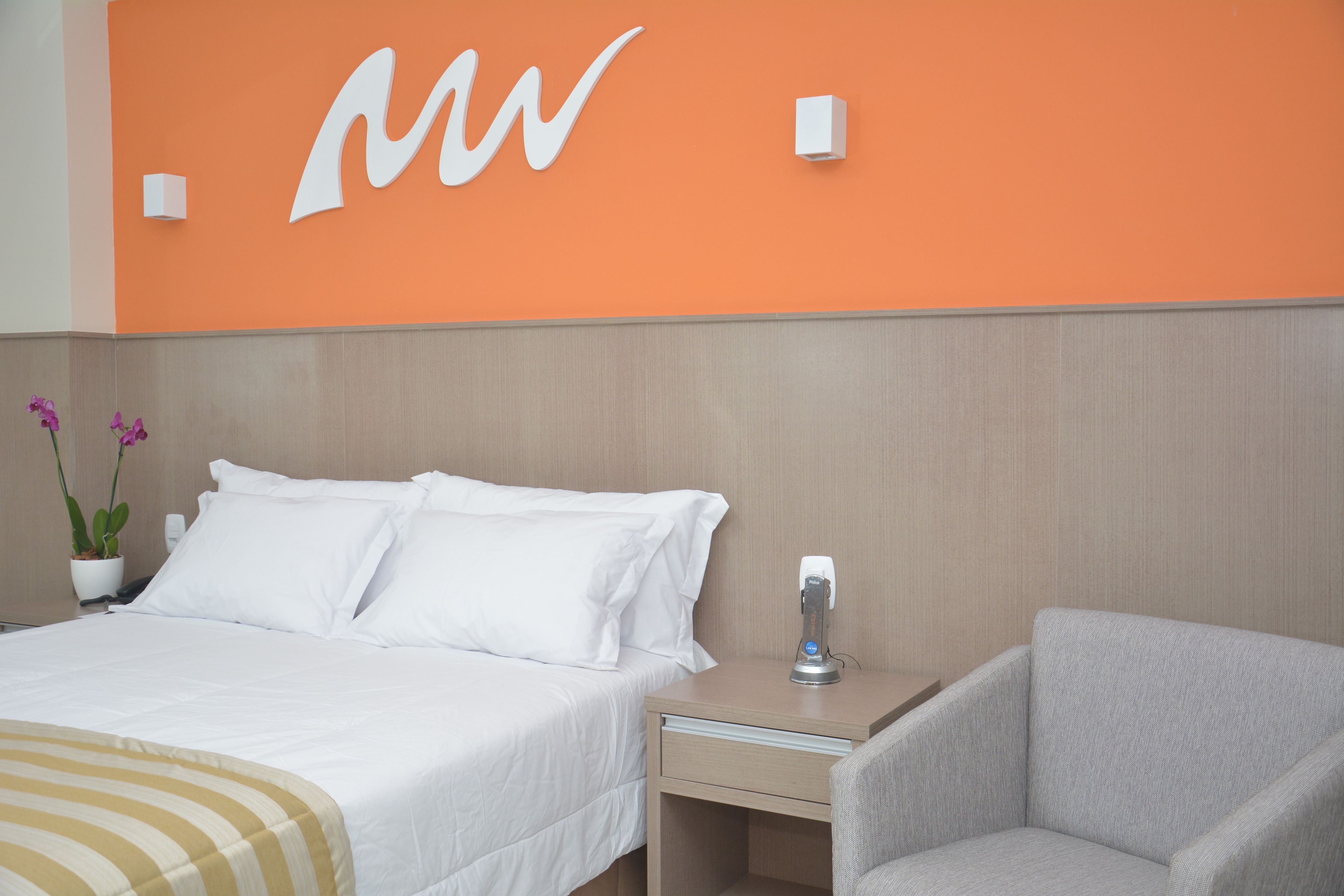 Habitación de hotel con cama, mesita de noche, una silla y pared con un gran logotipo 'M'.
