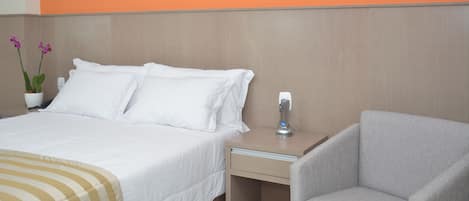 A hotel room with a bed, a nightstand, a chair, and a wall with a large 'M' logo.