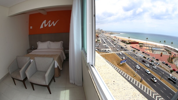 Quarto de hotel com cama, duas poltronas e vista para uma rua movimentada e praia.