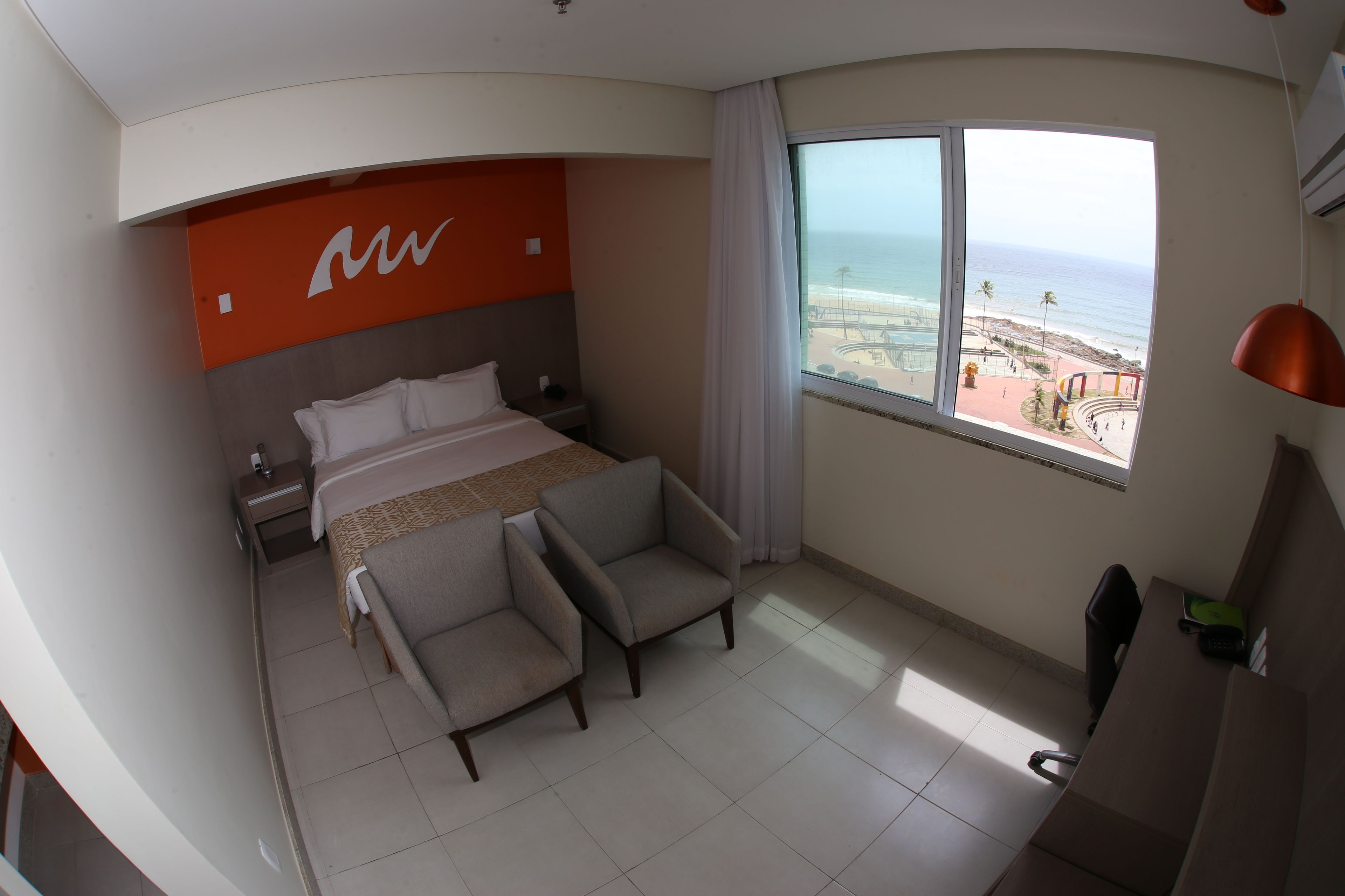 Ein Hotelzimmer mit einem Bett, zwei Sesseln, einem Schreibtisch und einem großen Fenster mit Blick auf Strand und Meer.