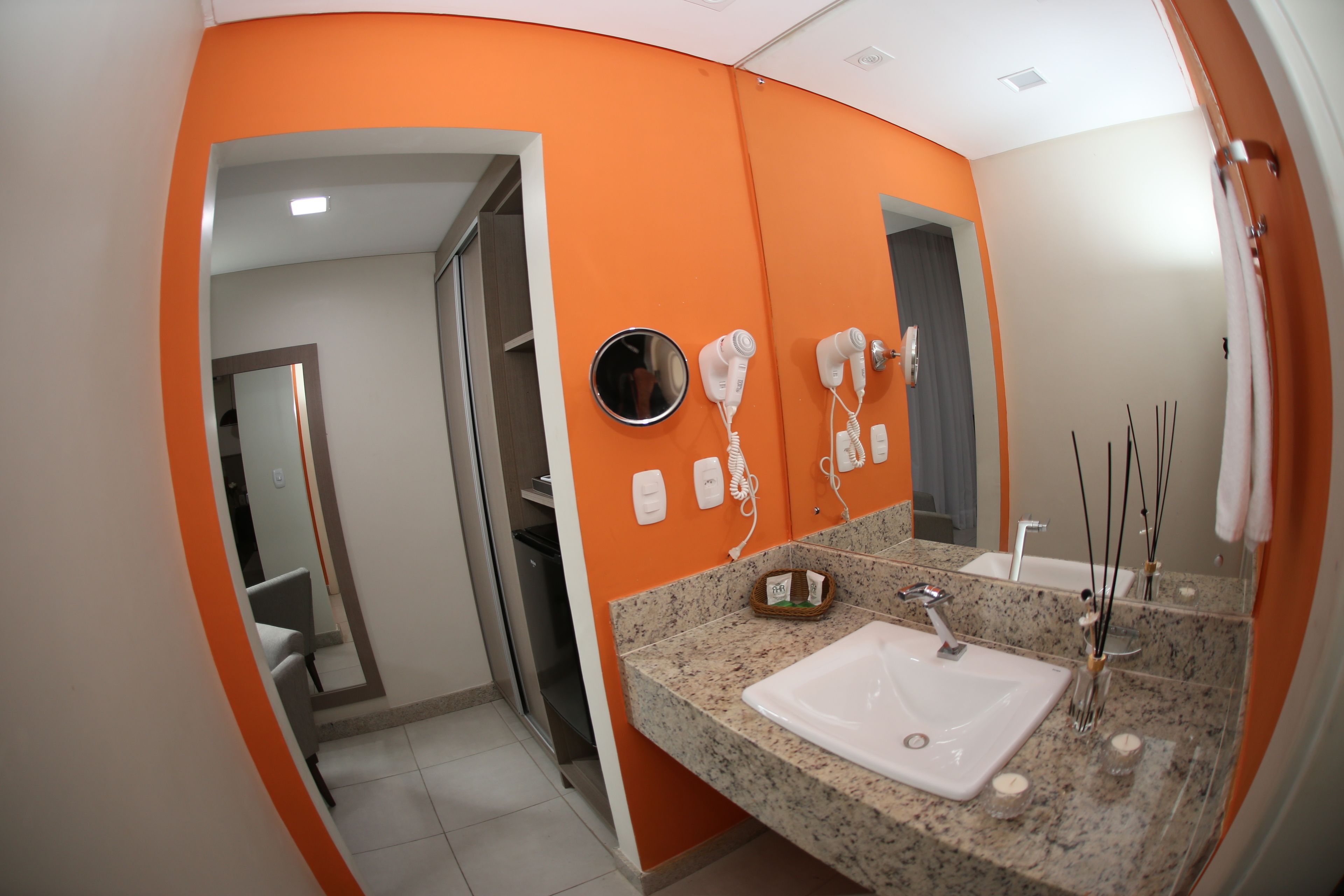 A modern bathroom with an orange accent wall, a granite countertop, a sink, a mirror, and two hair dryers mounted on the wall.