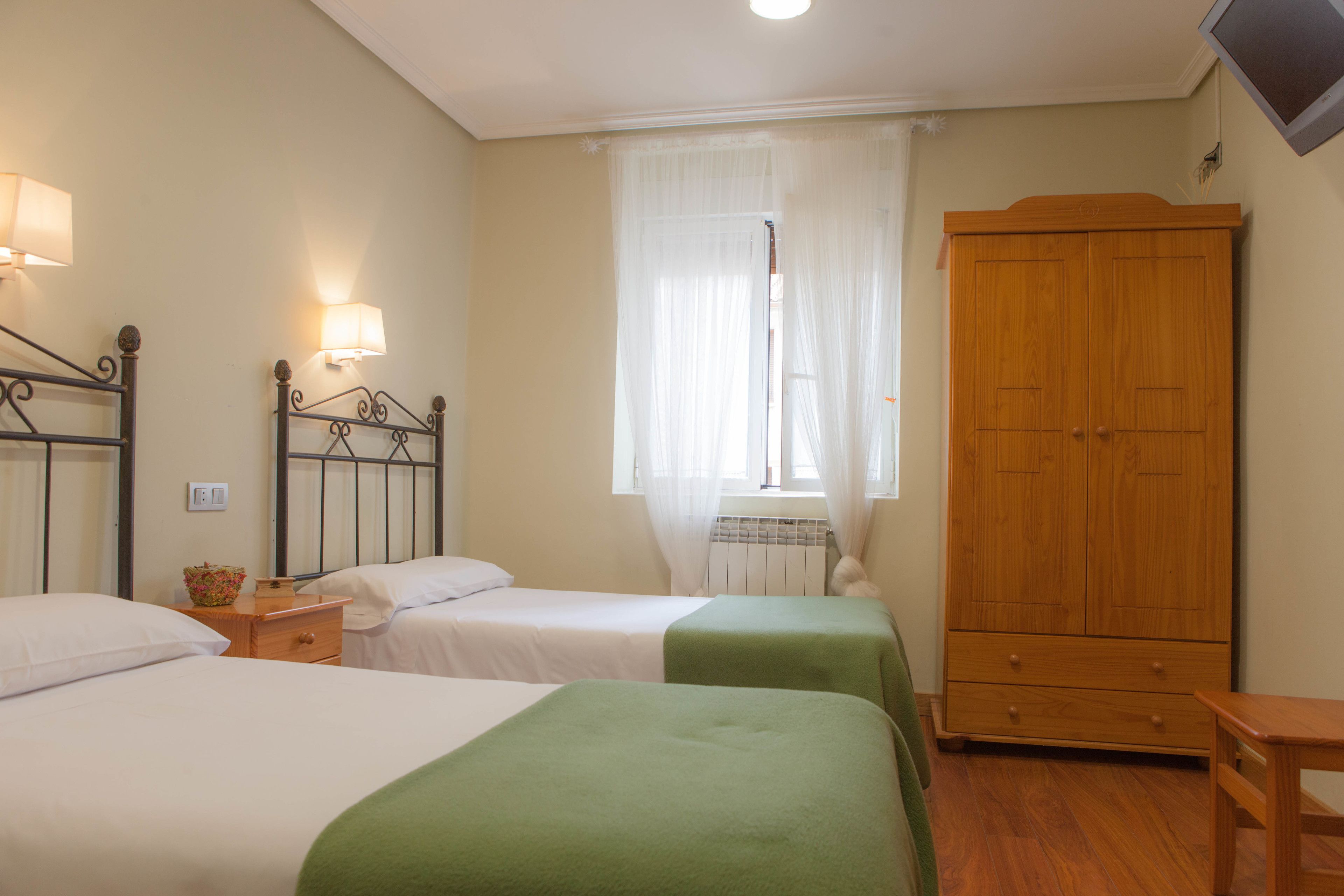 A bedroom with two beds, a wooden wardrobe, a bedside table, and a window with sheer curtains.