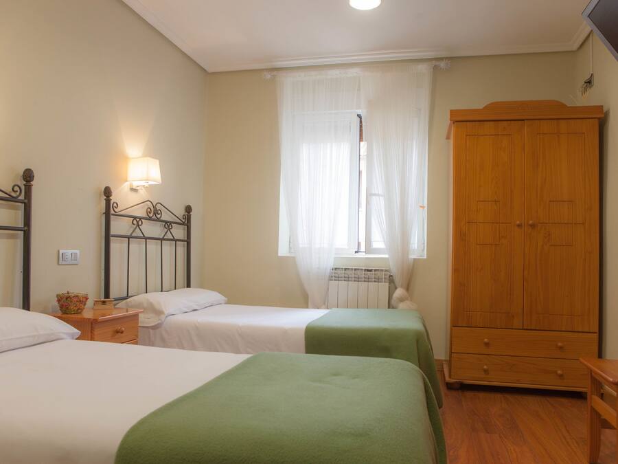 A bedroom with two beds, a wooden wardrobe, a bedside table, and a window with sheer curtains.