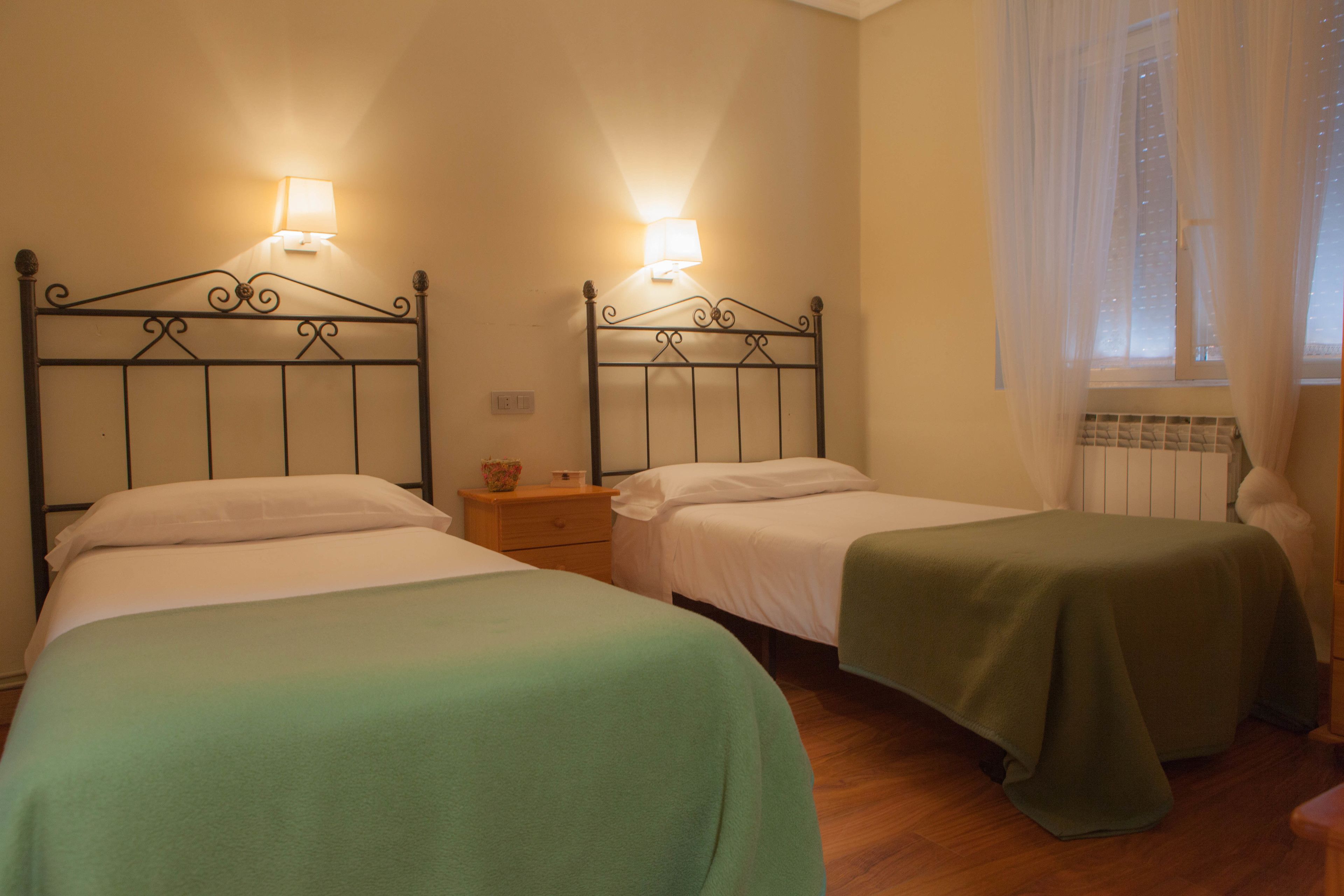 Two single beds with metal frames, each with a bedside table and a lamp. The room has wooden flooring and a window with sheer curtains.
