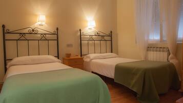 Two single beds with metal frames, each with a bedside table and a lamp. The room has wooden flooring and a window with sheer curtains.