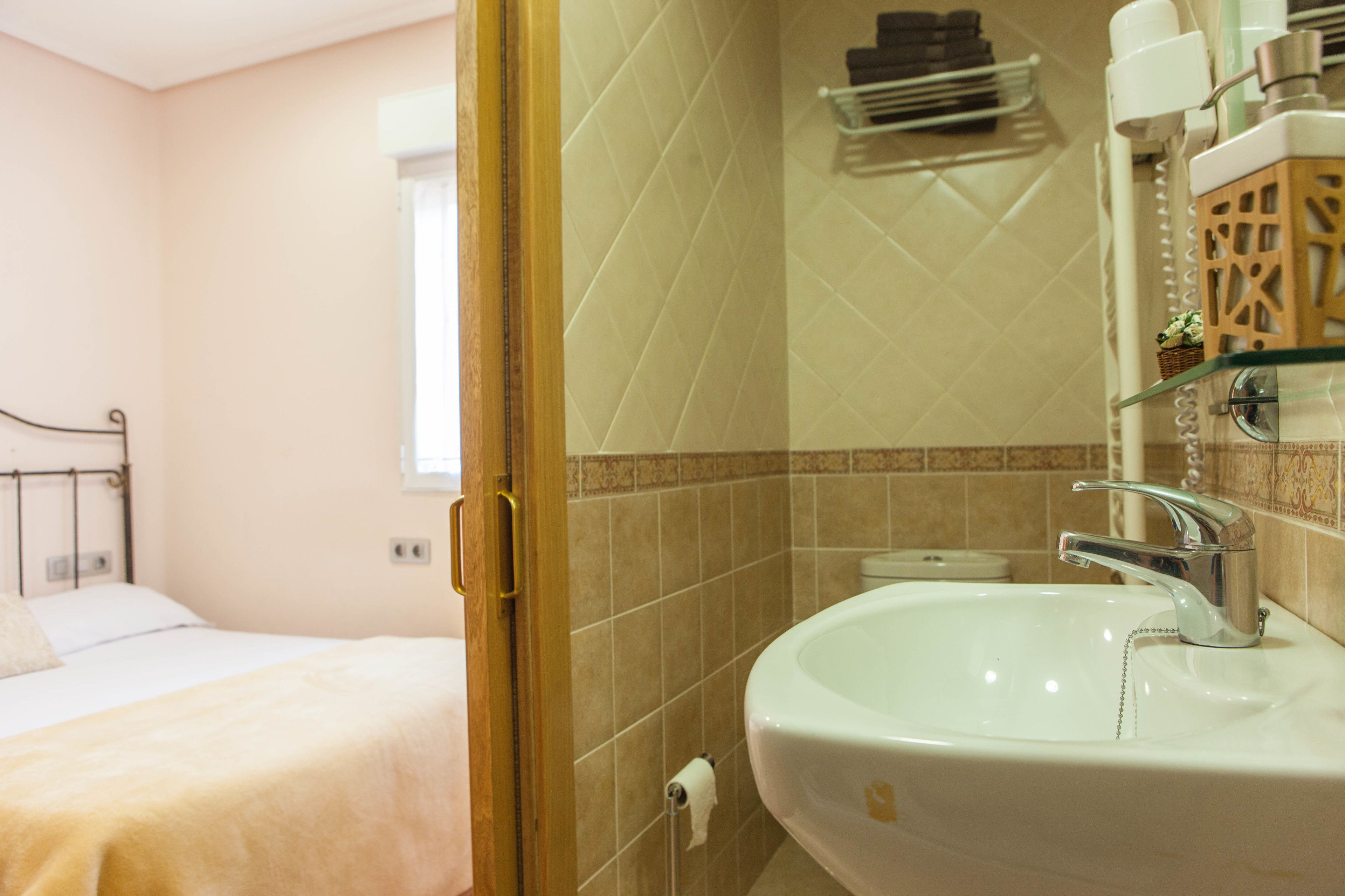 A hotel bathroom with a white sink, a glass shelf, and a bed in the adjacent room.