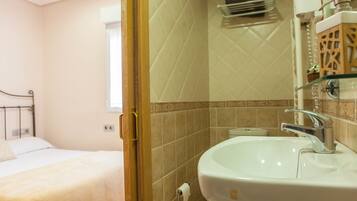 A hotel bathroom with a white sink, a glass shelf, and a bed in the adjacent room.