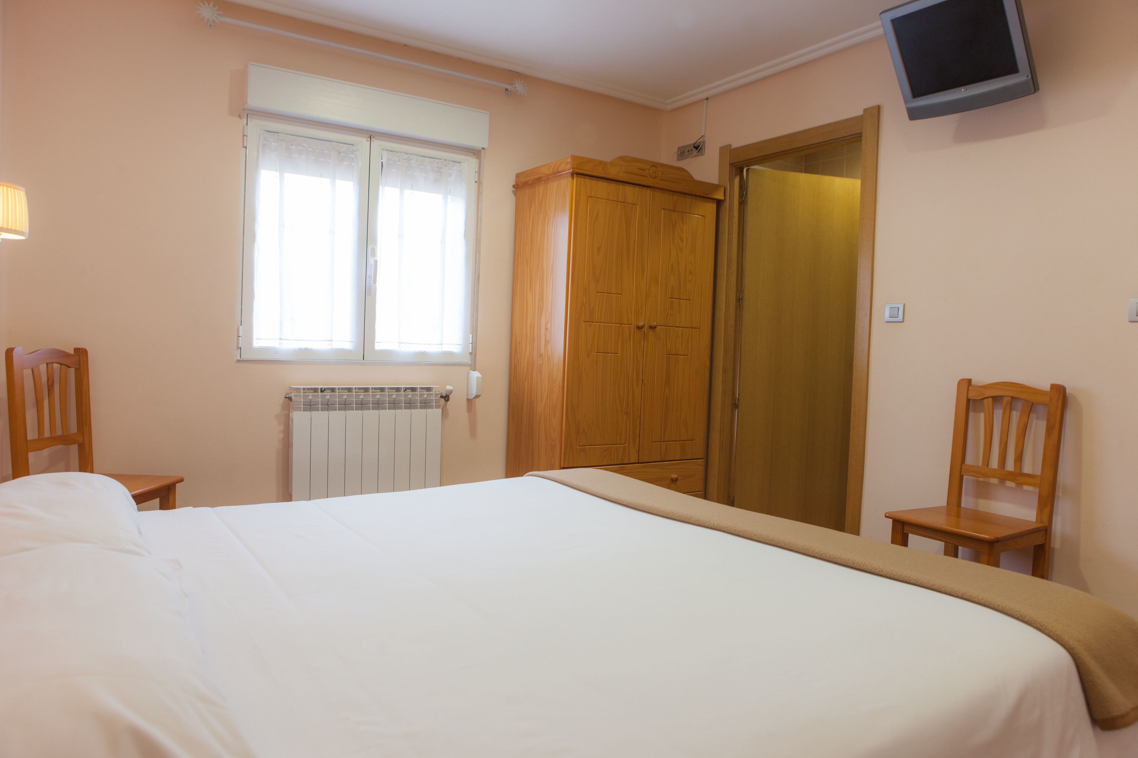 A bedroom with a bed, wooden wardrobe, a radiator, a window with curtains, and a television on the wall.