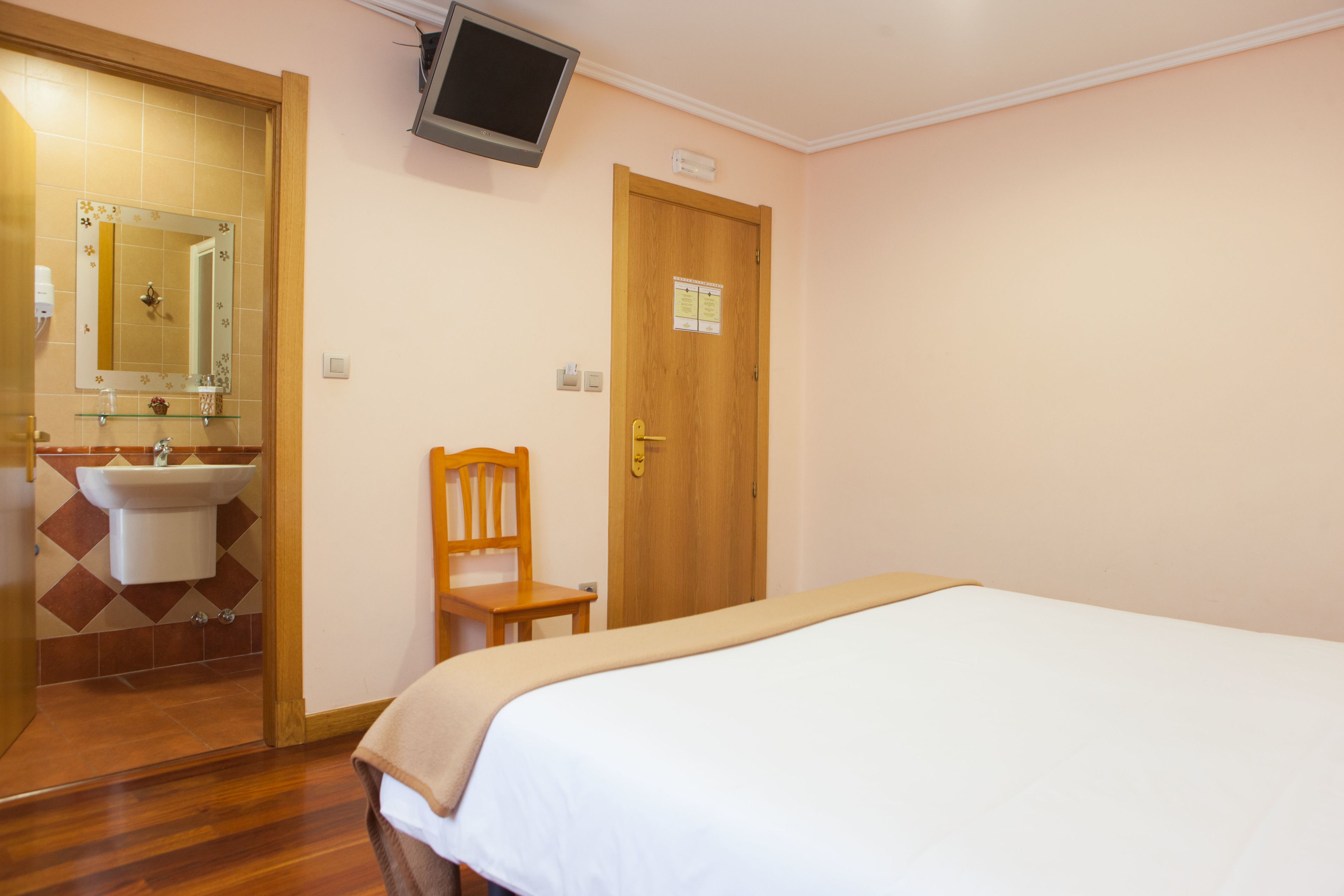 Un dormitorio con cama, una silla de madera, una puerta de madera, televisor montado en la pared y un baño con lavamanos y espejo.