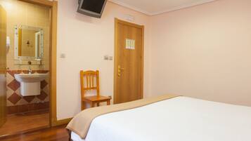 Un dormitorio con cama, una silla de madera, una puerta de madera, televisor montado en la pared y un baño con lavamanos y espejo.