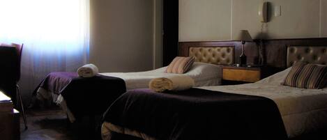 A hotel room with two beds, a wooden floor, a nightstand with a lamp, and a window with curtains.