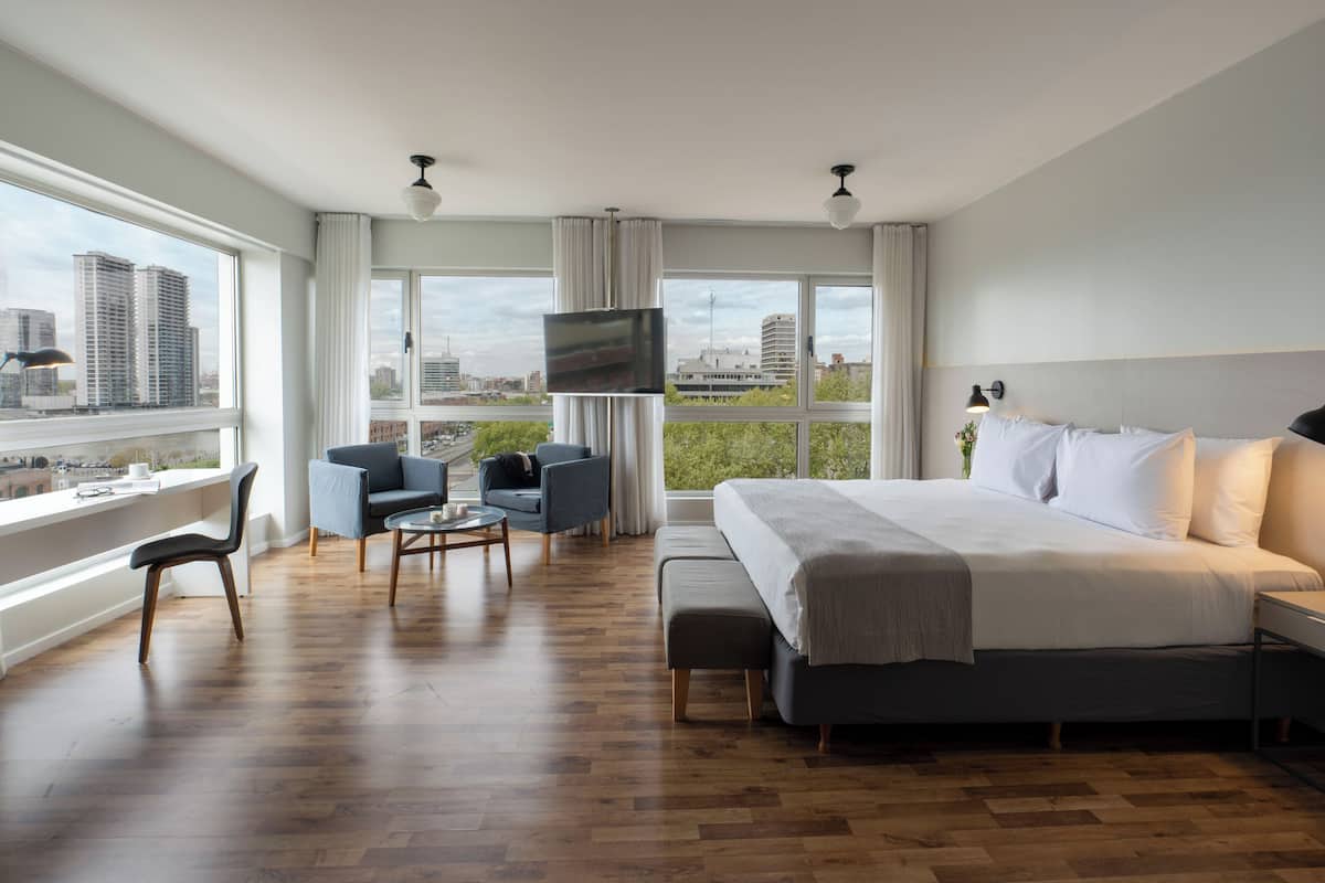 A hotel room with a large bed, a TV, a seating area with chairs, and a view of the city.