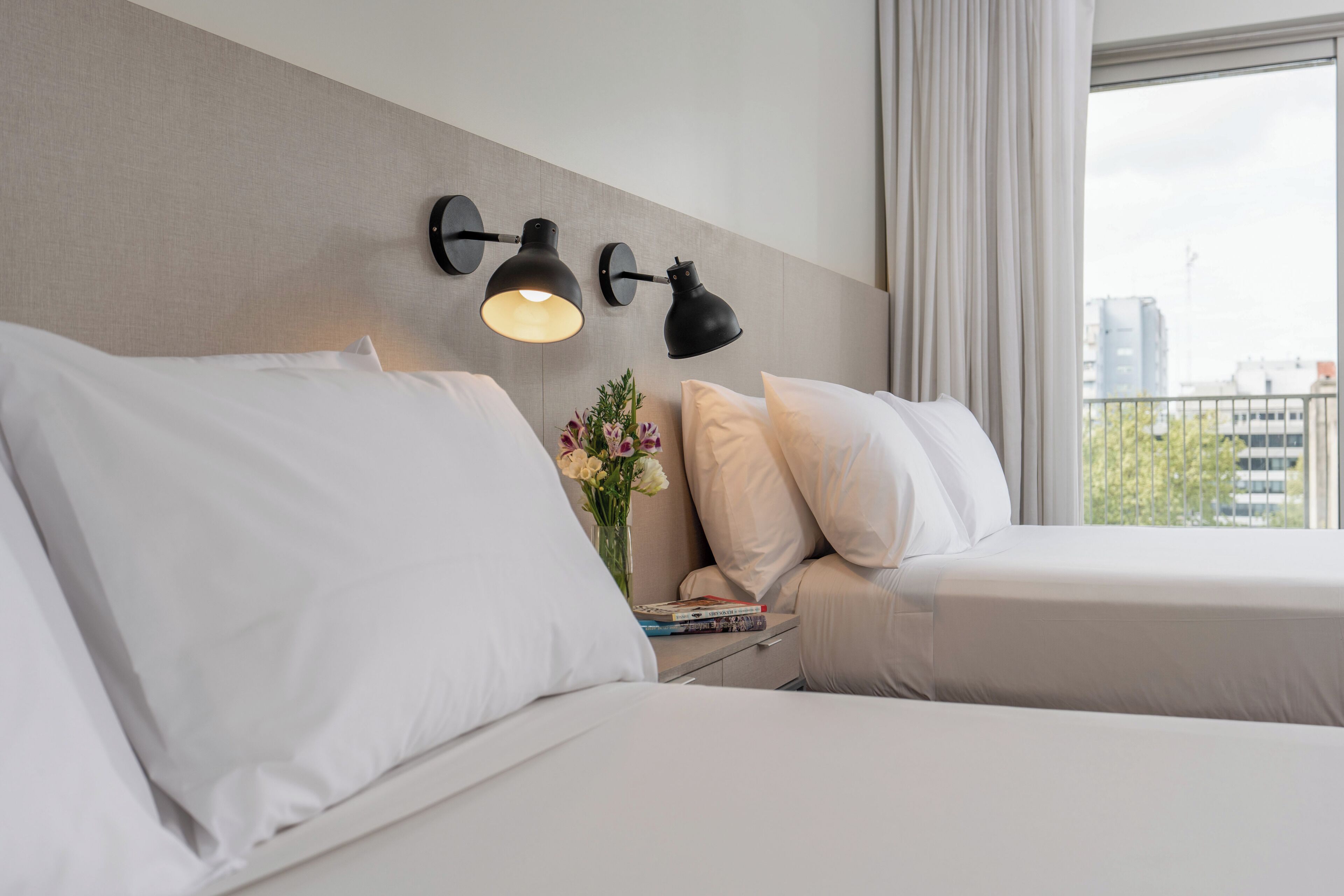 A hotel room with two beds, a nightstand with a vase of flowers, and a view of the city through the window.