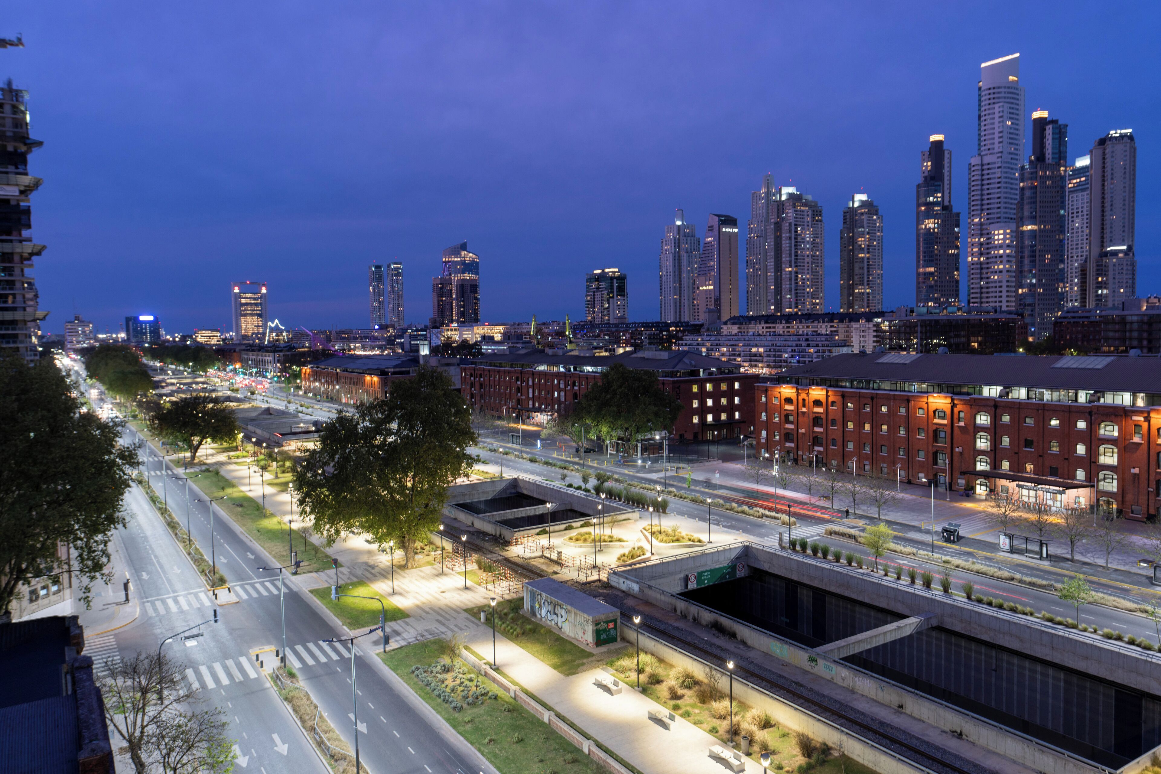 Un paisaje urbano al anochecer con modernos rascacielos, una intersección bien iluminada y una combinación de edificios comerciales y residenciales.